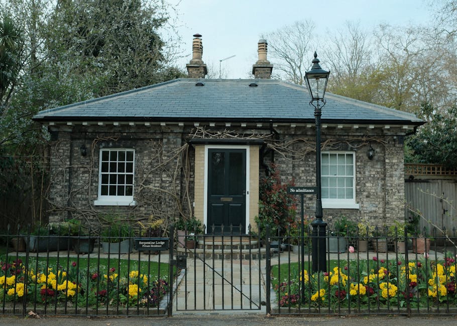 A small, charming brick cottage with two white-framed sash windows and a central black door, set behind a black wrought-iron fence with a small gate. The garden in front features neatly trimmed green 