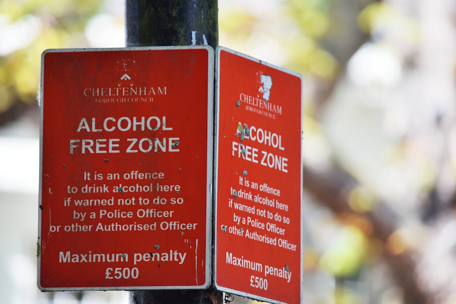 Two adjacent red and white signs mounted on a black metal pole in a garden setting, indicating an alcohol-free zone under Cheltenham Borough Council regulations. The signs feature white text stating '