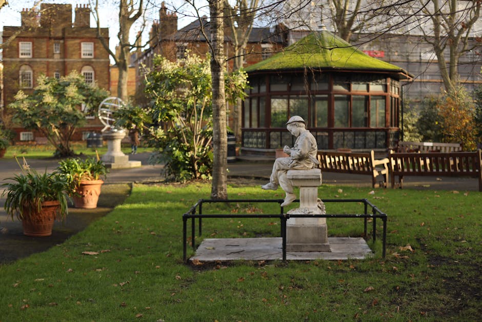 Two adjacent red and white signs mounted on a black metal pole in a garden setting, indicating an alcohol-free zone under Cheltenham Borough Council regulations. The signs feature white text stating '