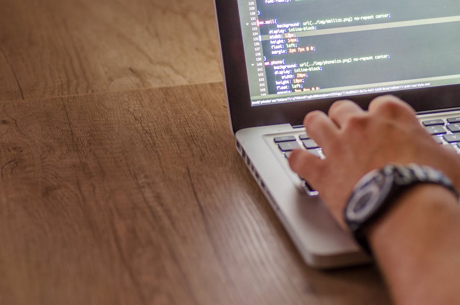 The image shows a person's right hand with a wristwatch working on a laptop placed on a wooden surface. The laptop screen displays lines of code with various colors, indicating programming or web deve